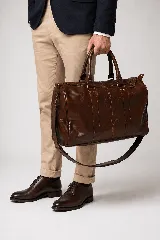 Bolso de viaje color marrón, confeccionado en cuero vacuno, con doble asa de mano y correa ajustable y desmontable. Cuenta con herrajes metálicos y costuras reforzadas.