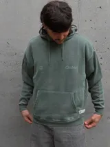Canguro con capucha de color verde con efecto lavado, diseño de corte holgado, bolsillo frontal tipo canguro, cordones ajustables y logo estampado en el pecho.