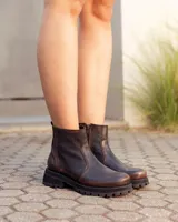 Bota de caña alta confeccionada en cuero color marrón caoba con acabado desgastado. Presenta una suela track robusta, correas con hebillas en la parte superior de la caña y un arnés decorativo con herrajes metálicos en el tobillo.