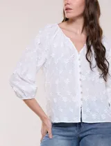 Blusa blanca de calce holgado, con bordado en relieve tipo broderie de motivos florales en toda la prenda. Presenta escote en V con fruncido en los hombros, cierre frontal con botonadura y mangas tres cuartos abullonadas con puños elastizados.
