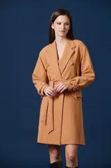 Saco largo tipo trench de color tostado, con diseño de doble botonadura, solapas clásicas y cinturón ajustable en la cintura. Presenta mangas largas con trabillas en los puños y una silueta estructurada y fluida.