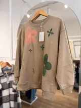 Buzo de cuello redondo en color beige, con diseño oversize y felpa interna. Presenta bordados de flores en tonos rosa, verde y azul distribuidos en el frente.