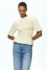 Remera de punto de algodón color amarillo claro, con cuello redondo, hombros levemente caídos, mangas cortas y cuerpo completamente fruncido con detalle de volados en el ruedo.