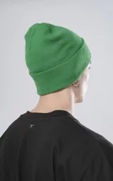 Gorro de tejido de punto color verde, de estilo beanie con borde doblado y un pequeño parche negro con un logo blanco de tijeras cruzadas.