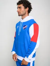 Canguro de felpa Umbro del Club Nacional de Football, con diseño tricolor en rojo, azul y blanco. Presenta capucha con cordones ajustables, bolsillo tipo canguro frontal, logo de la marca y escudo del club bordados en el pecho.