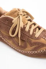 Zapatilla de perfil bajo confeccionada en gamuza combinada en tonos marrones. Presenta detalles de flecos en el lateral, costuras decorativas en forma de X a lo largo de la base y cordones al tono.