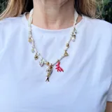 Collar corto hecho con cinta de tela blanca y dijes dorados, rojos y nacarados con forma de pez, corazón, coral, flor y cuentas.