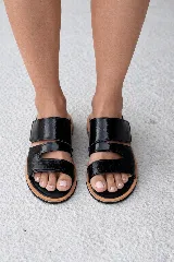 Sandalia plana de cuero color negro con dos tiras anchas ajustables mediante velcro. La tira superior es lisa y brillante, mientras que la tira inferior tiene textura de cocodrilo. La plantilla es acolchada y forrada en cuero de color claro, con una suela de goma de doble capa en negro y marrón claro.