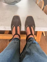 Ballerinas de gamuza color marrón oscuro con diseño tipo T-strap y detalles de costuras decorativas en color verde oliva. Cuentan con cierre de pulsera al tobillo y suela plana.