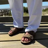 Sandalia tipo Birkenstock con doble tira ajustable de cuero sintético negro y hebillas metálicas. Cuenta con plantilla anatómica de corcho y suela de goma.