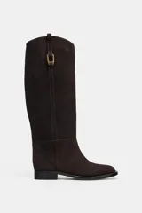 Bota de caña alta confeccionada en serraje, con diseño de estilo hípico. Presenta detalle de pespuntes verticales, tira decorativa con hebilla metálica en la parte superior de la caña y puntera redonda. Cuenta con suela plana de 2,5 cm y sistema de calce tipo pull-on.
