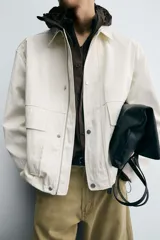 Cazadora de hombre, corte relaxed fit, color blanco hueso o crema, con acabado brillante. Presenta cuello solapa, manga larga, dos bolsillos de plastrón con solapa en la cadera y cierre frontal con botones a presión ocultos por una solapa.