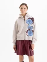 Buzo con cierre frontal y capucha, color beige, con estampado floral y de cadenas en el frente. La capucha cuenta con forro de corderito al tono, mangas largas con puños elásticos y ajuste holgado.
