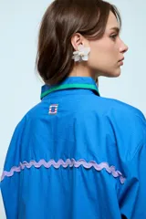 Camisa azul de corte oversize, con cierre frontal de botones y detalles de cintas onduladas en colores contrastantes a lo largo de la botonadura y en los puños. Presenta cuello clásico y mangas largas.