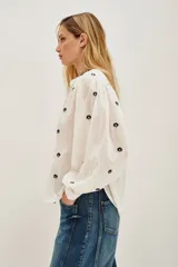 Camisa de algodón color crudo con bordados florales pequeños en azul marino. Presenta mangas largas abullonadas, cuello redondo y una abertura frontal con botones y detalles de encaje calado en la pechera y hombros.