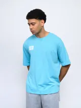 Remera de corte holgado color celeste, con cuello redondo diferenciado y logo de la marca estampado en el pecho. Presenta un diseño de manga corta y estilo deportivo.