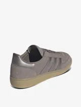 Championes Adidas modelo Handball Spezial de perfil bajo, confeccionados en gamuza color marrón topo con las clásicas tres tiras y detalles metalizados en tono plateado, inscripción lateral en relieve y suela de caucho translúcido.