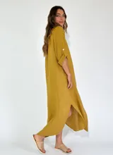 Vestido camisero largo de lino, color mostaza, con cuello camisero, cierre frontal de botones y mangas tres cuartos con detalle de tejido calado en los laterales.