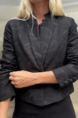 Blazer corto tipo spencer, color negro, con cuello mao y tejido con bordado tono sobre tono en patrón paisley. Presenta cierre frontal y botones forrados en las mangas.