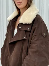 Campera de gamuza color marrón con cuello de corderito sintético en color crema. Presenta un diseño oversize con cierre frontal, solapas con broches metálicos y una tira ajustable en el ruedo.