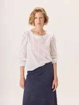 Camisa blanca de algodón con diseño de broderie, cuello redondo y mangas largas con volados en los hombros y puños elásticos.