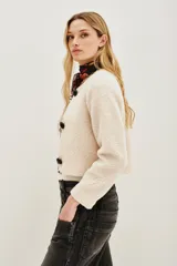 Saco corto de tejido tipo boucle en color crudo, con cierre frontal mediante tres trabas de cuero sintético negro y botones esféricos. Presenta ribetes en contraste negro a lo largo de la abertura frontal y el cuello redondo.