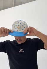 Gorro tipo jockey color beige con lunares azules, visera azul y bordado frontal con la palabra "ABOIU" en letras de colores y estrellas.