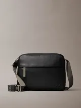 Bolso bandolera rectangular de Calvin Klein, color negro, con textura de cuero granulado. Posee un bolsillo frontal horizontal con cierre y el logo CK grabado en la parte inferior derecha. La correa es ancha y de tela en tono gris/verde oliva, ajustable con hebilla metálica.