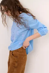 Blusa celeste de manga larga con delicados bordados florales en tono más claro en la zona del pecho y mangas. Presenta escote en V y puños elásticos abotonados.
