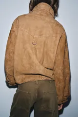 Cazadora de ante 100% color tostado, con cuello alto ajustable mediante hebilla, cierre frontal de cremallera metálica, mangas largas con trabillas ajustables en los puños y bajo elástico.