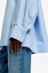 Camisa oversize de algodón color celeste, con cuello camisero, botones frontales y mangas largas ranglán con puños abotonados. Presenta pespuntes a contraste en toda la prenda.