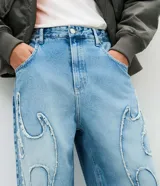 Pantalón de jean corte super baggy, color azul medio, con aplicaciones tribales y recortes desflecados. Cuenta con cintura con pasadores, cierre frontal con botón y cremallera, y bolsillos funcionales.