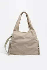 Bolso de hombro Bimba y Lola de gran tamaño, confeccionado en nylon color beige. Presenta una estructura blanda, doble asa de mano, correa regulable extraíble y bolsillos laterales con cierre. Incluye logo metálico de la marca en la parte inferior frontal.