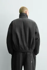 Cazadora de corte relajado en color gris oscuro con diseño de rayas diplomáticas. Presenta cuello solapa, cierre frontal con cremallera, bolsillos laterales de vivo y puños elásticos. Incluye un detalle de texto bordado en contraste en la parte frontal.