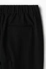 Pantalón negro de tejido suave con corte relajado, cintura elástica con cordón oculto y pliegues frontales. Presenta bolsillos laterales y bolsillos traseros ribeteados.