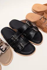 Sandalias de cuero color negro con dos tiras anchas sobre el empeine, cada una con hebillas metálicas ajustables y detalles de tachas plateadas. La suela es de cuero con un borde de color madera.