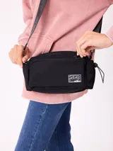 Riñonera negra de diseño rectangular con cierre frontal y compartimento principal. Presenta una etiqueta rectangular con el logo de la marca en el frente y correa ajustable con el nombre de la marca estampado.