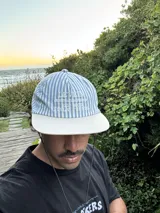 Gorro tipo gorra de visera plana, con paneles a rayas verticales azules y blancas, y visera blanca lisa. Presenta bordado en la parte frontal con el texto "LEVEIN", "MOOD THAT ONLY KNOW" y "SEE GOOD FIALN THINGS".