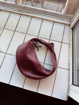 Cartera tipo hobo de cuero color borgoña con asa superior anudada.