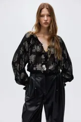 Blusa de corte holgado en tejido jacquard transparente con diseño de hojas metalizadas. Presenta cuello mao con frunces, abertura frontal con tiras finas, ribete de volados y mangas largas tipo globo con puños elásticos.