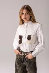 Camisa blanca de manga larga con cuello alto gorguera con volados y detalles de alforzas verticales en el frente. Cuenta con dos bolsillos en el pecho de pelo con estampado de vaca en tonos marrones y blancos.