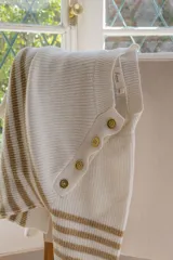 Sweater de punto en color crema con rayas horizontales en tono dorado. Presenta un diseño de cuello redondo con una solapa lateral decorativa que incluye botones dorados. De corte holgado y manga larga.