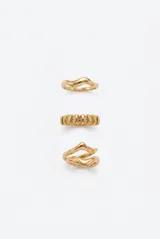 Set de tres anillos metálicos dorados con diseños irregulares y orgánicos, incluyendo un modelo ondulado, uno con textura de barras verticales y uno con forma de doble banda entrelazada.