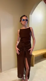 Vestido largo de color marrón con diseño de escote recto y tirantes finos. Presenta un detalle de nudo frontal en la cintura que crea un efecto drapeado y una abertura central pronunciada que deja ver una capa inferior más corta.