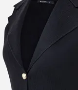 Chaleco negro de tejido punto, sin mangas, con cuello solapa y cierre frontal de dos botones dorados. El corte es acinturado y alargado.