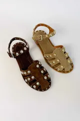 Sandalias planas de gamuza color tostado, con múltiples tiras decoradas con tachas metálicas doradas semiesféricas. Cuenta con una tira ajustable en el tobillo con hebilla metálica.