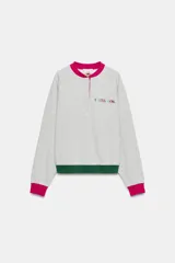 Sudadera gris melange de la colección especial Fruit of the Loom x Zara, con cuello redondo fucsia con abertura y botones, mangas largas con puños fucsias, y ribete inferior verde oscuro. Cuenta con logo bordado multicolor en el pecho.
