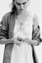 Vestido midi color beige de viscosa, sin mangas, con escote en V profundo adelante y atrás decorado con ribetes de encaje. Presenta una falda acampanada con bajo asimétrico y costuras en zigzag.