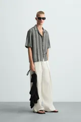 Camisa de calce relajado confeccionada en tejido jacquard con estructura abierta y diseño geométrico. Presenta cuello bowling, manga corta, aberturas laterales en el bajo y cierre frontal con botones.