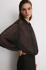 Cazadora semitransparente de color negro, con capucha, cierre frontal de cremallera metálica y mangas largas abullonadas con puños elásticos. Presenta un diseño holgado con bajo elástico.